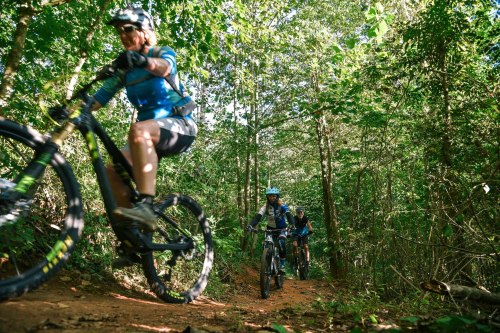 A group of mountain bikers riding on a dirt trail through a lush green forest.