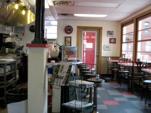 Cozy diner interior with tables, chairs, and a counter; large windows let in natural light.
