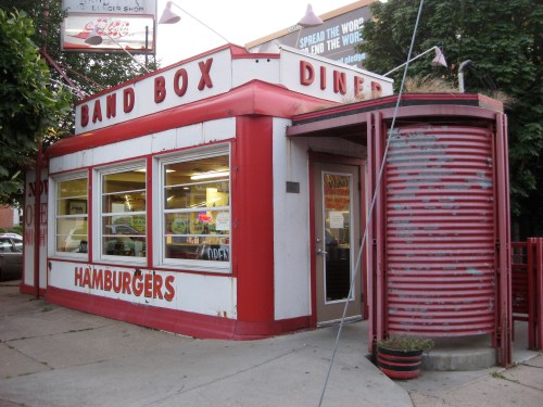 A retro diner with a red and white exterior, featuring a sign that reads "Band Box Hamburgers" and large windows.