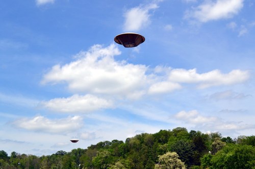 Two flying saucers hover in a blue sky above a green landscape with trees and fluffy white clouds.