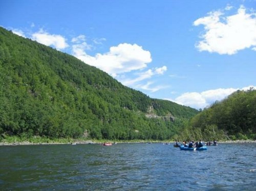 A scenic river surrounded by lush green mountains under a clear blue sky, with people rafting in the water.