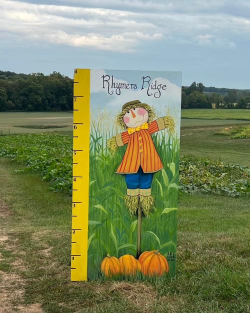A colorful scarecrow painted on a height chart stands in a field with pumpkins and green crops in the background.