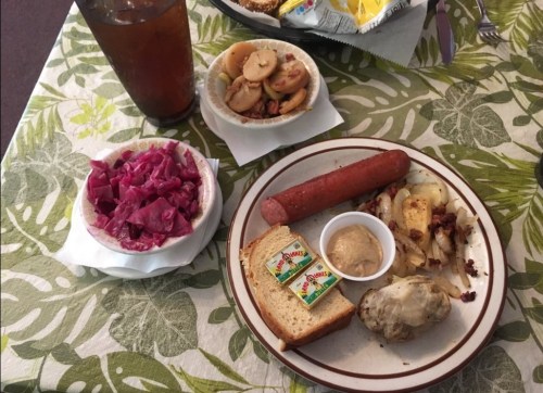 A plate with a sausage, bread, mustard, artichoke, and sides of red cabbage and fruit, with a drink in the background.