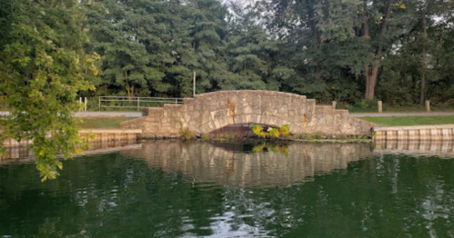 A stone bridge arches over a calm, green-tinted pond surrounded by trees.