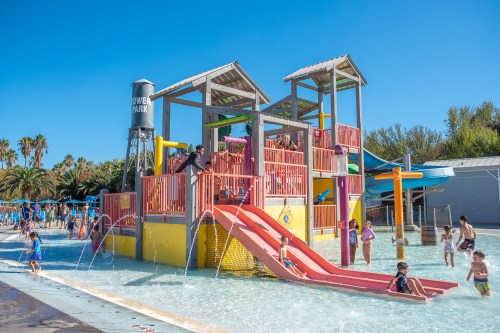 Colorful water play structure with slides and kids playing in a sunny water park setting. Palm trees in the background.