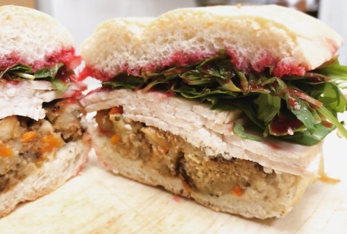 Close-up of a sandwich with layers of turkey, stuffing, greens, and cranberry sauce on soft bread.