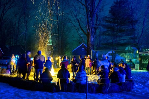 A group of people gathered around a campfire at night, with sparks rising against a dark, snowy backdrop.