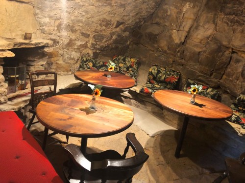 Cozy underground café with stone walls, wooden tables, and floral cushions, featuring small vases of sunflowers.