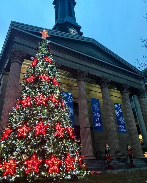 A brightly lit Christmas tree with red stars stands in front of a historic building adorned with festive banners.