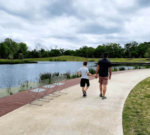 A man and a boy walk hand in hand along a path by a lake, with green trees and cloudy skies in the background.
