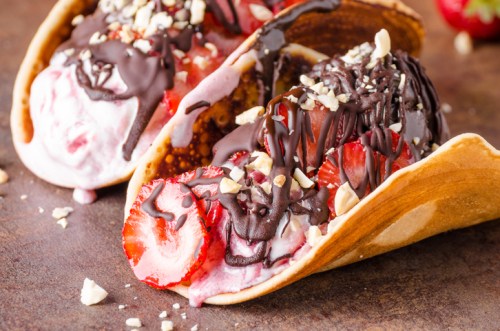 Two dessert tacos filled with ice cream, strawberries, chocolate drizzle, and chopped nuts on a rustic surface.