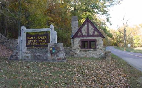 Sign for Sam A. Baker State Park with a stone structure and trees in the background, along a road.