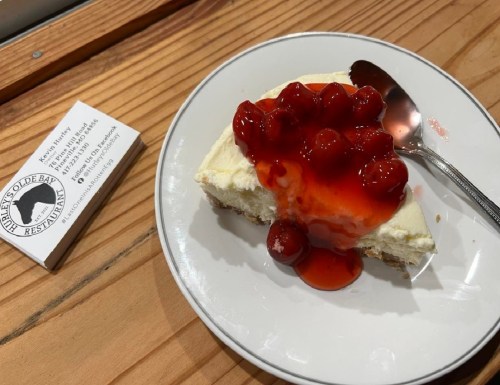 A slice of cheesecake topped with cherry sauce on a white plate, next to a business card on a wooden table.