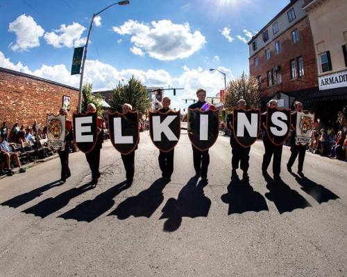 A parade scene with participants holding large signs spelling "ELKINS" against a sunny sky and crowd in the background.