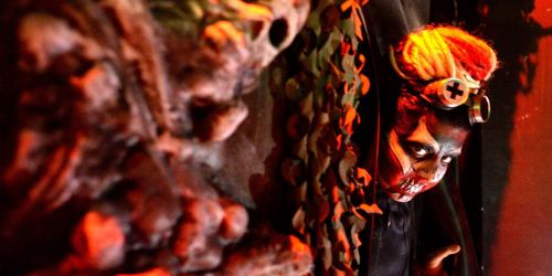 A person in a dark, eerie setting with dramatic lighting, wearing a creepy costume and makeup, peering from behind a wall.