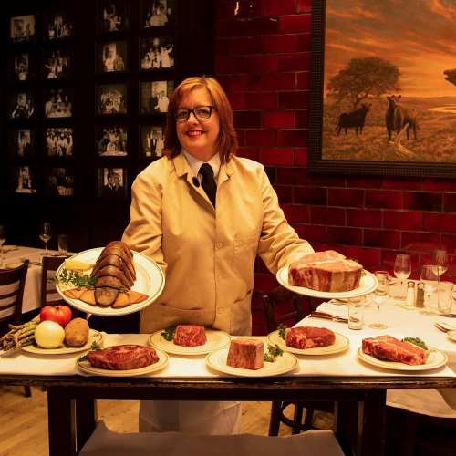 A smiling server in a beige jacket presents a variety of meats on plates at a restaurant, surrounded by elegant decor.