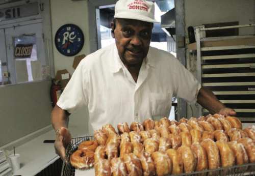 A man in a white shirt and cap holds a tray of glazed donuts in a bakery setting.