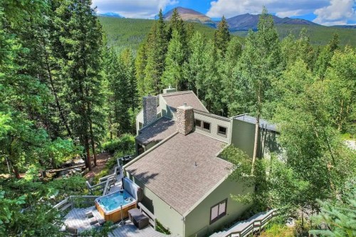 A cozy green cabin nestled among tall trees, featuring a hot tub on a wooden deck with mountains in the background.