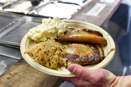 A hand holds a plate with grilled sausages, sauerkraut, and creamy potato salad.