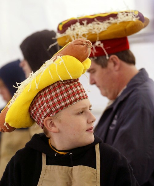 Two people wearing playful hot dog hats, one with a yellow and red hat and the other in the background.
