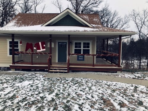 A cozy house with a porch decorated for the holidays, surrounded by snow and bare trees.