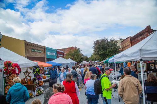 A bustling street fair with vendors under tents, crowds of people browsing, and colorful decorations on display.