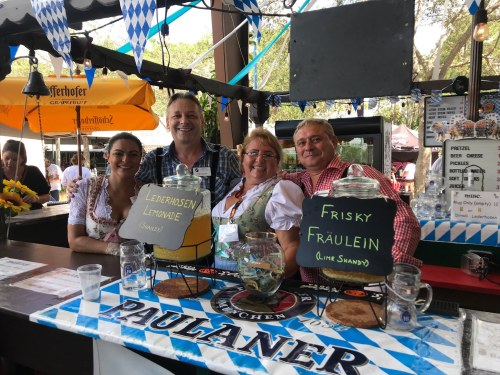 Four people in traditional attire stand behind a bar with drinks, smiling at the camera. Festive decorations are visible.