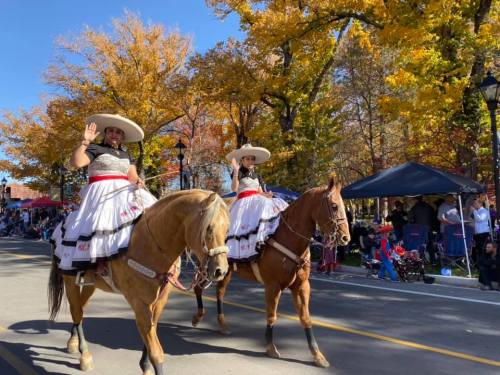 Two women in traditional dresses and wide-brimmed hats wave while riding horses in a festive parade with autumn trees.