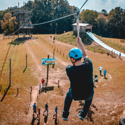 A person on a zip line glides over a grassy area with trees and a playground in the background.