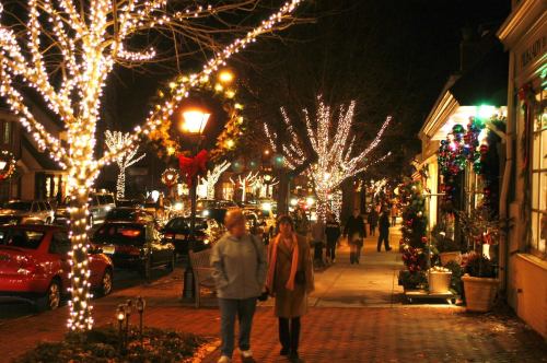 A festive street at night, adorned with holiday lights and decorations, with people walking along the sidewalk.