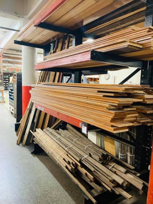 A storage area with wooden planks and boards stacked on shelves and the floor, in a warehouse setting.