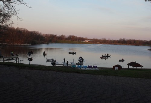 Boats on a calm lake at sunset, with trees in the background and a picnic area in the foreground.