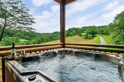A hot tub on a wooden deck overlooks a scenic landscape with rolling hills and a winding road.