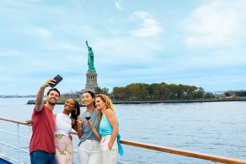 A group of four friends takes a selfie with the Statue of Liberty in the background on a boat.