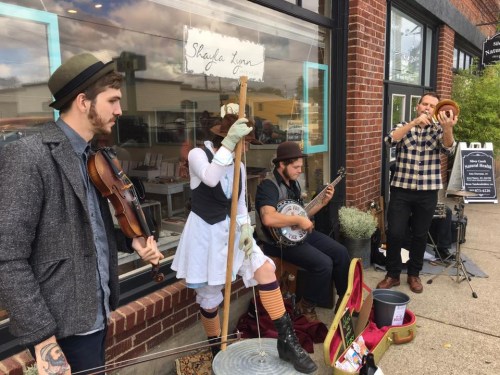 A street performance featuring four musicians: a fiddler, a banjo player, a flutist, and a performer holding a sign.