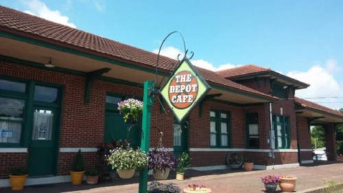 A brick building with a sign reading "The Depot Café" and colorful flower pots in front under a clear blue sky.