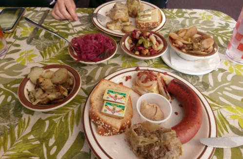 A table with two plates of food featuring sausage, bread, potatoes, and various sides like pickled vegetables and salad.