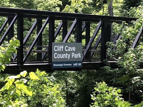 Sign for Cliff Cave County Park on a bridge surrounded by lush green trees.