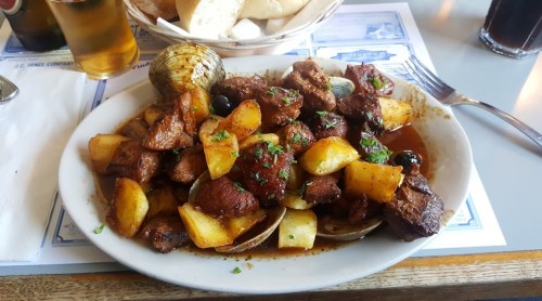 A plate of seasoned meat and potatoes, garnished with herbs, served with clams and a side of bread.