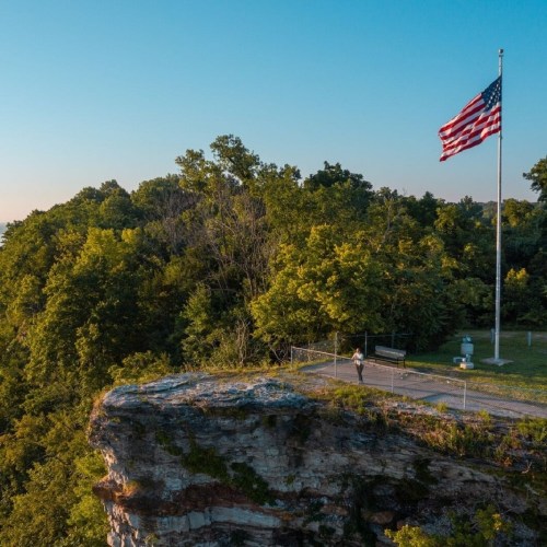 A scenic view from a cliff overlooking lush green trees and a flagpole, with a person standing on a viewing platform.