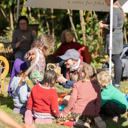 A group of children and adults engage in a hands-on activity outdoors, surrounded by greenery and colorful materials.