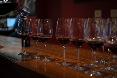 A hand pours red wine from a carafe into a row of empty wine glasses on a wooden bar.