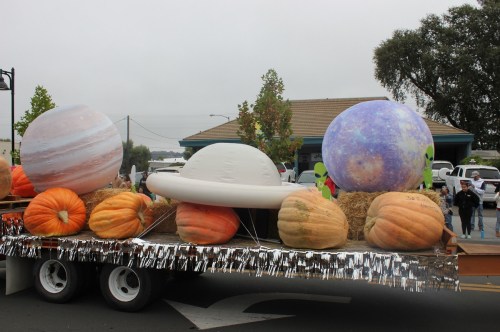 A parade float featuring large planet models and pumpkins, with aliens and hay bales, set in a festive atmosphere.