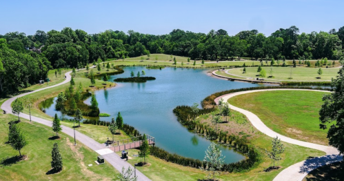 A serene park with a winding pond, lush greenery, and walking paths under a clear blue sky.