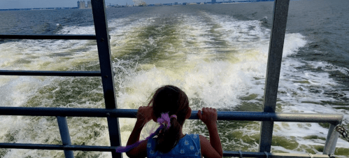 A child with a ponytail looks out over the water from a boat, watching the wake created by the vessel.