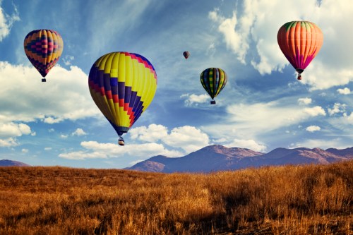 Colorful hot air balloons float over a golden field with mountains and a blue sky in the background.