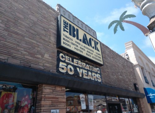 Sign for "The Black" store, celebrating 50 years, featuring tobacco, music, and accessories, with a palm tree in the background.