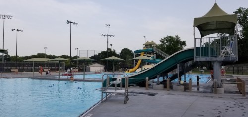 A swimming pool area featuring a water slide, lounge chairs, and people enjoying the water on a sunny day.