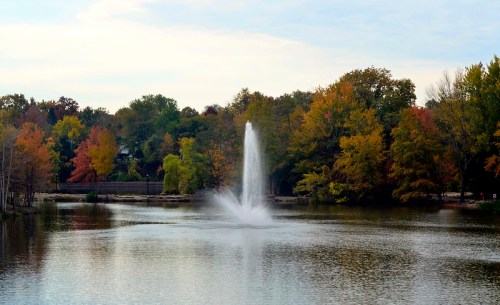 A serene lake with a fountain spraying water, surrounded by trees displaying autumn colors.