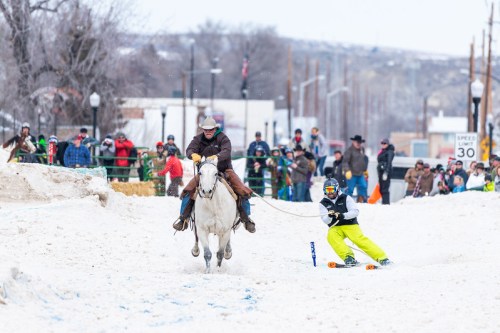 A cowboy on a horse pulls a skier through a snowy street, surrounded by a cheering crowd in winter attire.